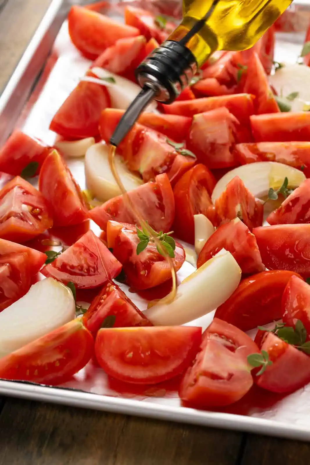 Olive oil being drizzled over cut tomatoes, onions, and herbs on a sheet pan.