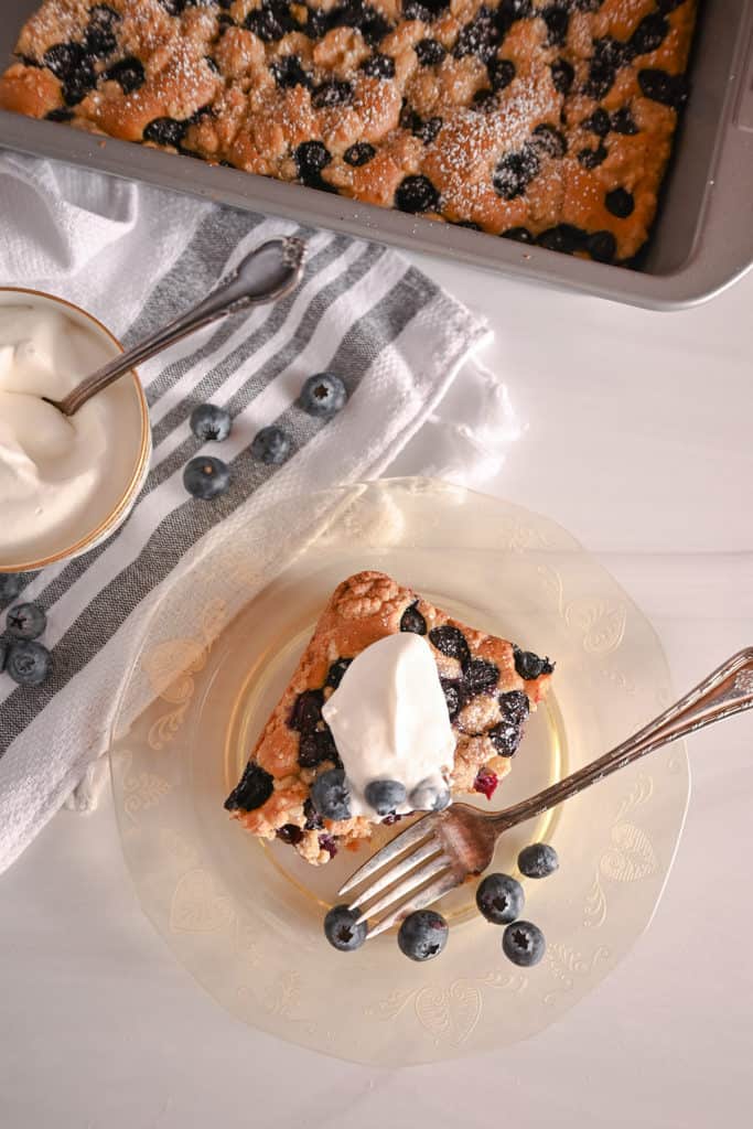 Overhead view of a slice of blueberry crumb cake on a plate, topped with whipped cream, with a bite taken from the corner