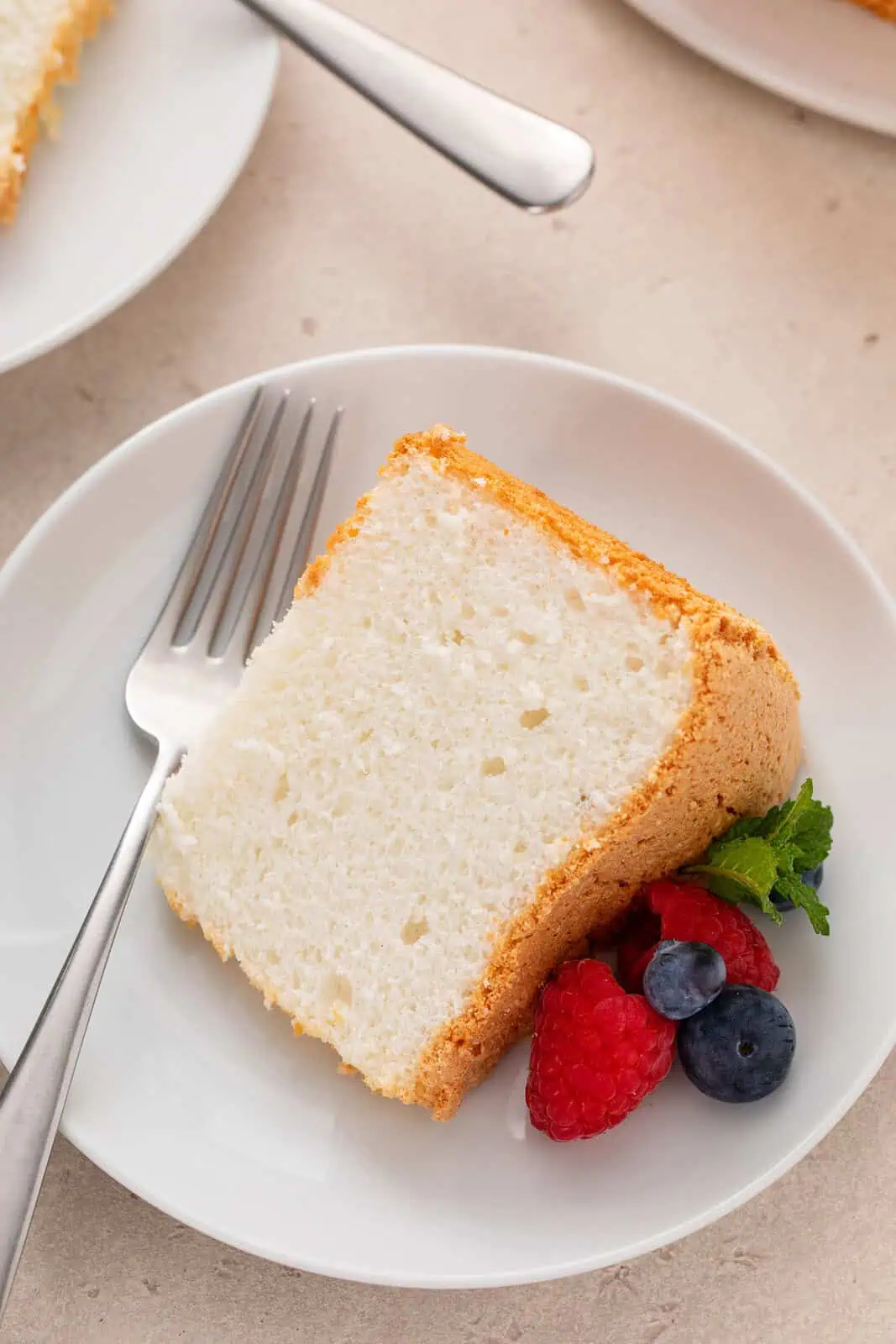 Close up of a slice of angel food cake on a white plate with a fork and fresh berries.