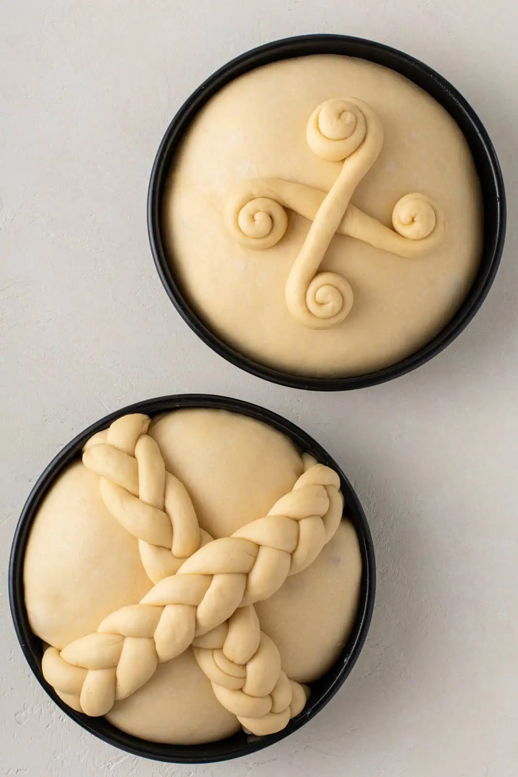 Risen shaped paska loaves in two round cake pans, topped with dough decorations. One loaf is topped with criss-crossed braids and the second is topped with swirls.