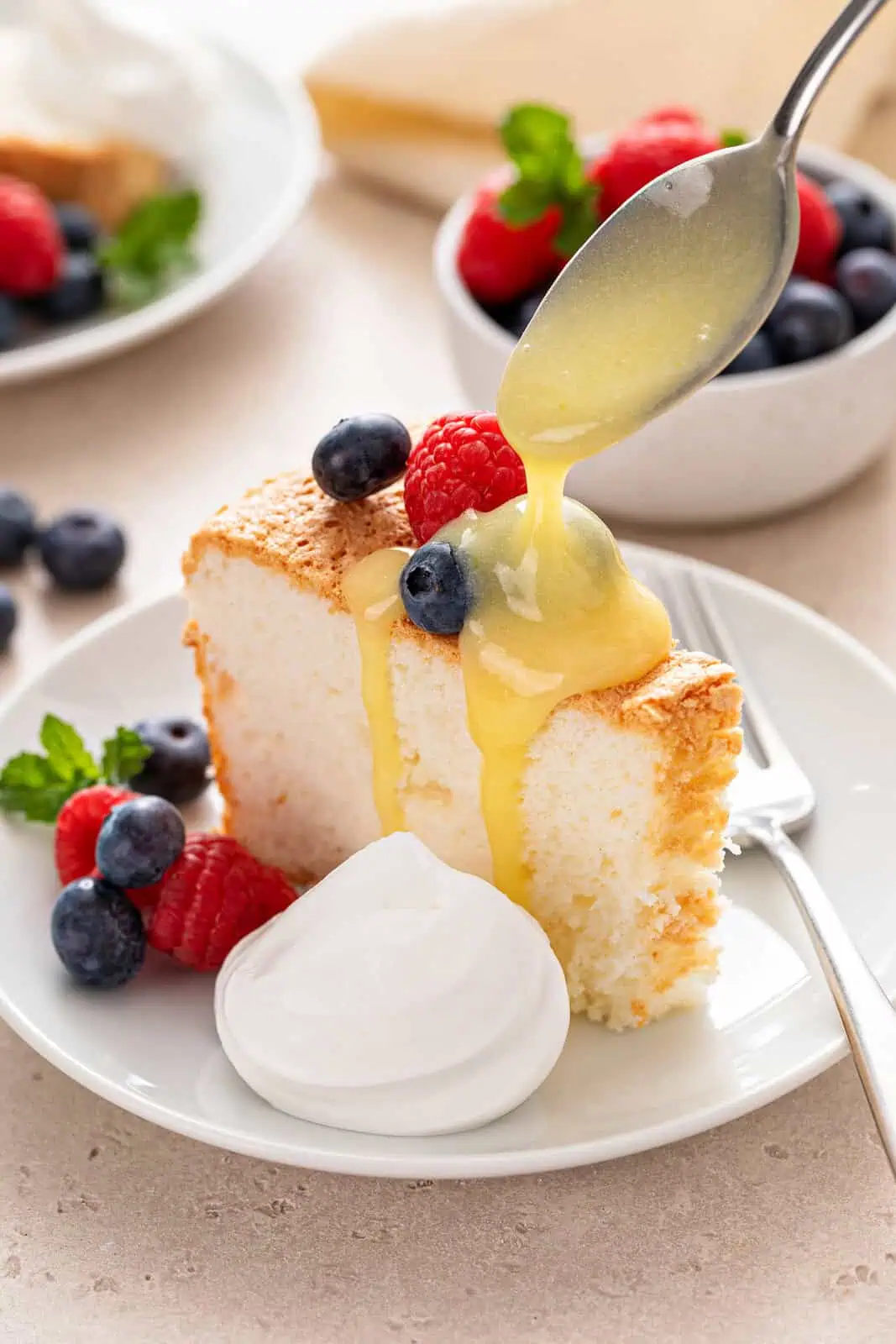 Lemon curd being drizzled over a slice of angel food cake on a white plate. The cake is also topped with fresh berries and set next to a dollop of whipped cream.
