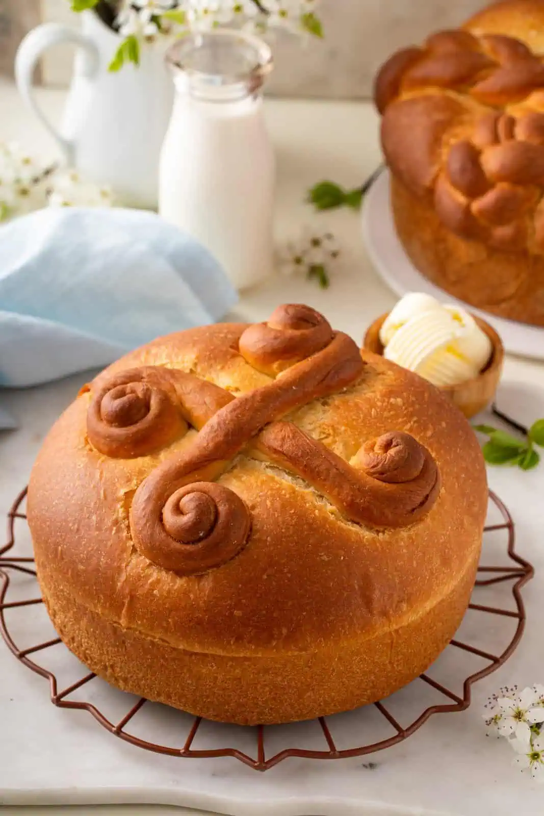 Cooled loaf of paska on a wire rack. a second loaf, a bowl of butter, and a small jug of milk are in the background.