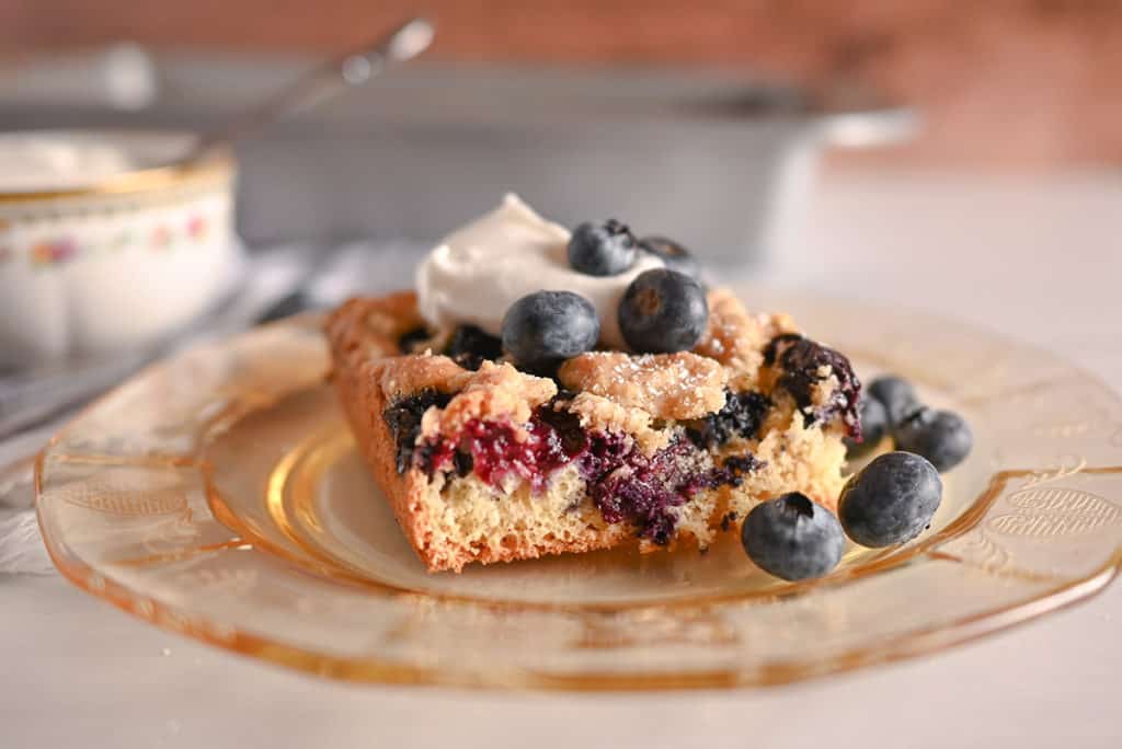 Side view of a plated slice of blueberry crumb cake topped with whipped cream and blueberries