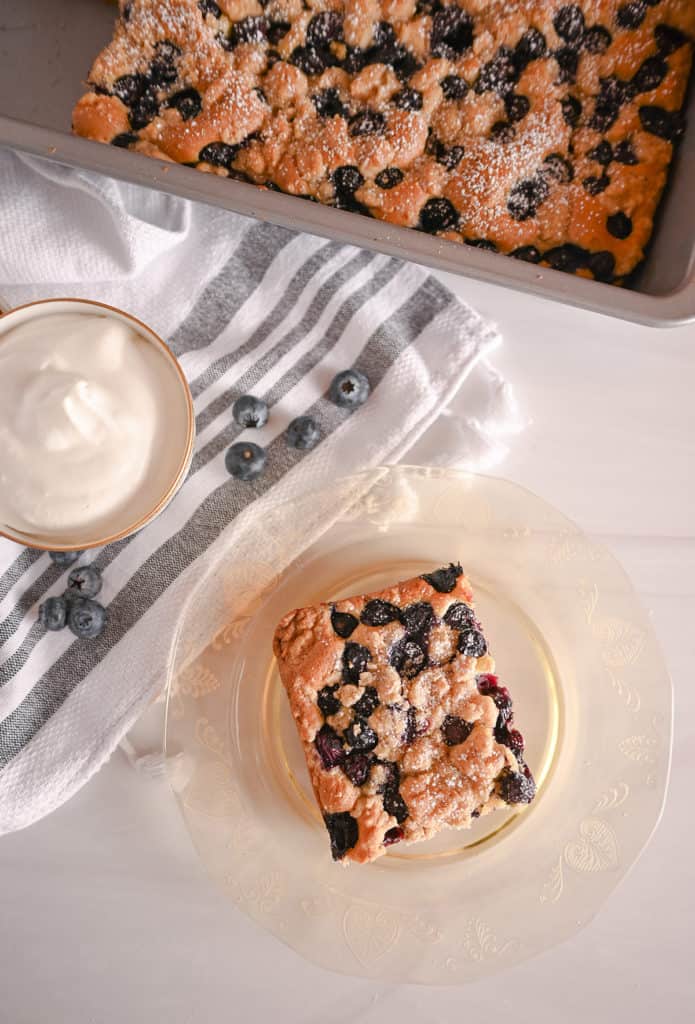 Overhead view of a slice of blueberry crumb cake on an amber plate, next to a bowl of whipped cream