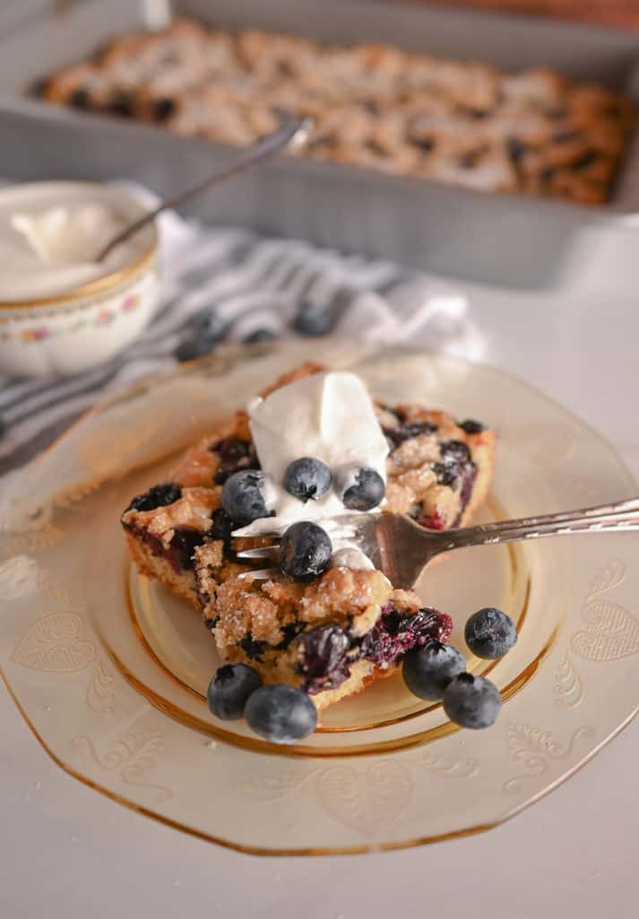 Fork cutting a bite from the corner of a slice of blueberry crumb cake plated on an amber plate