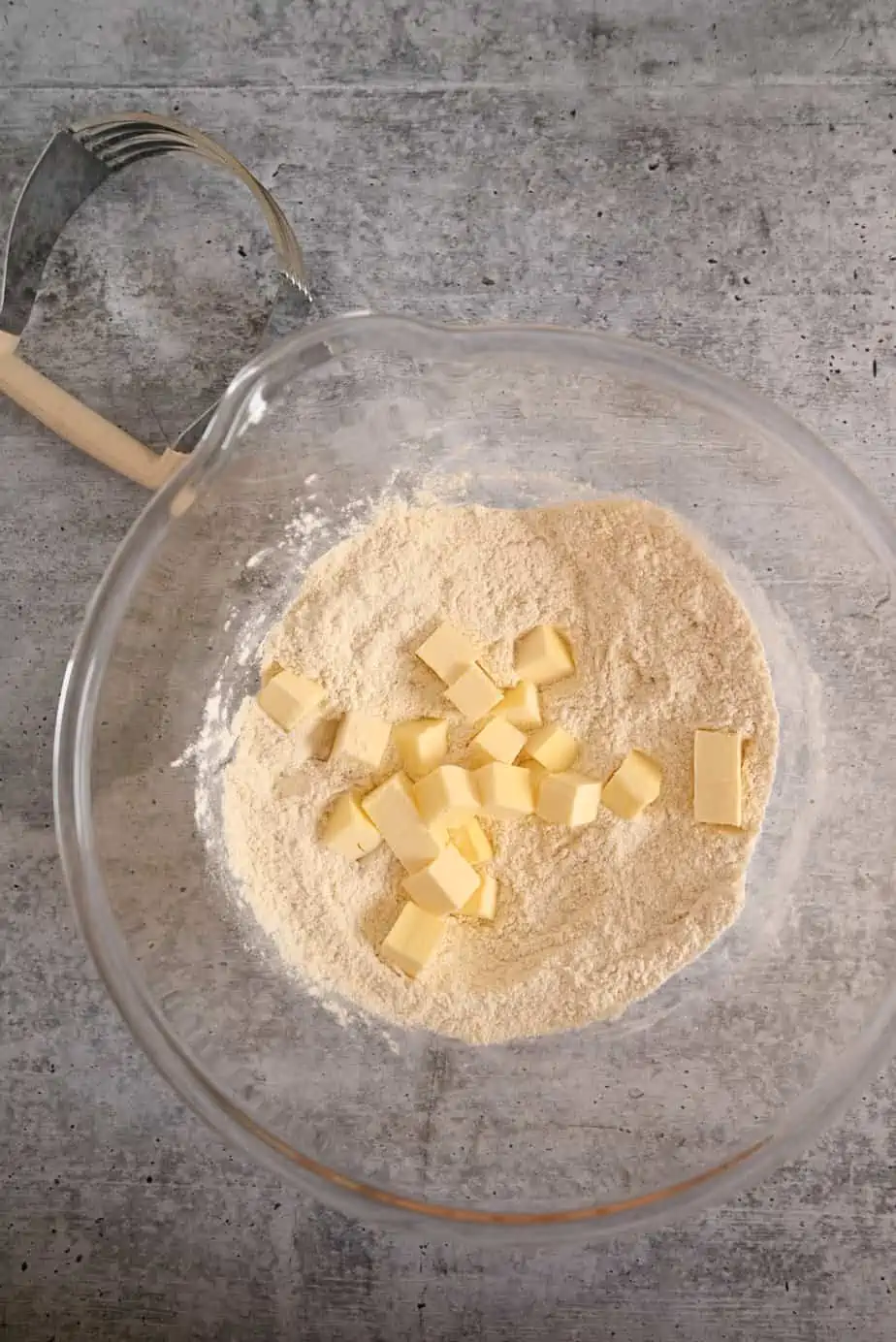 Cubed butter being added to dry ingredients in a glass mixing bowl.