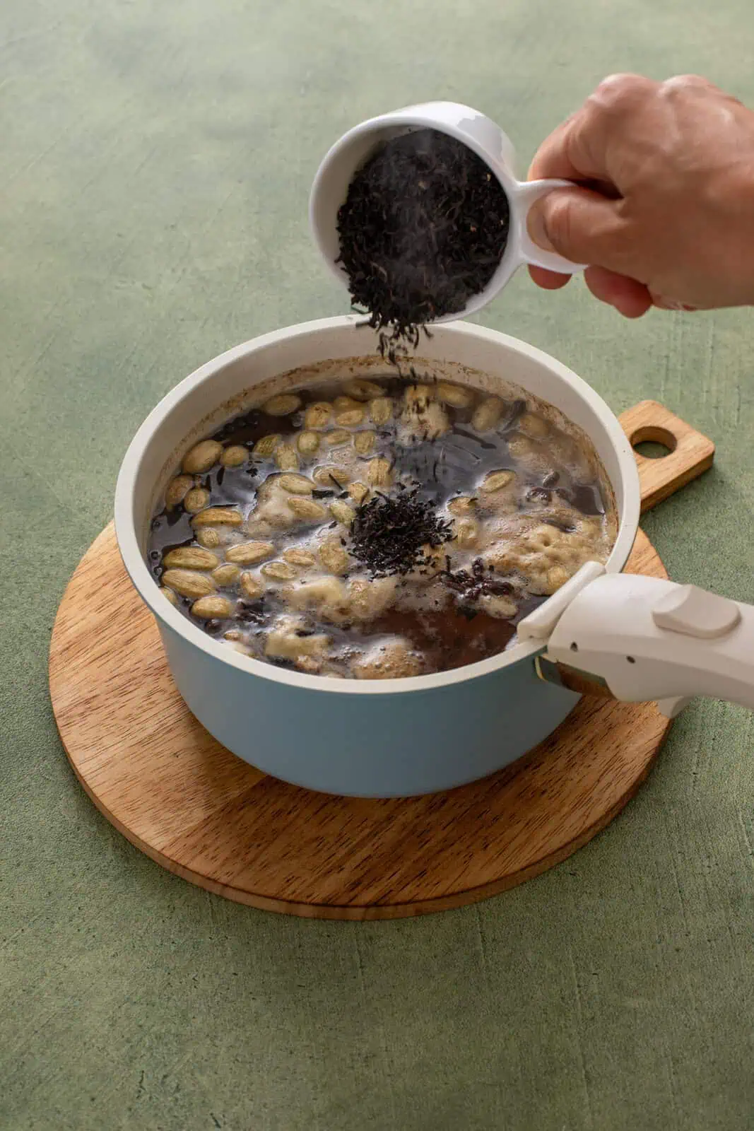 Person adding black tea leaves to chai concentrate base in a saucepot.