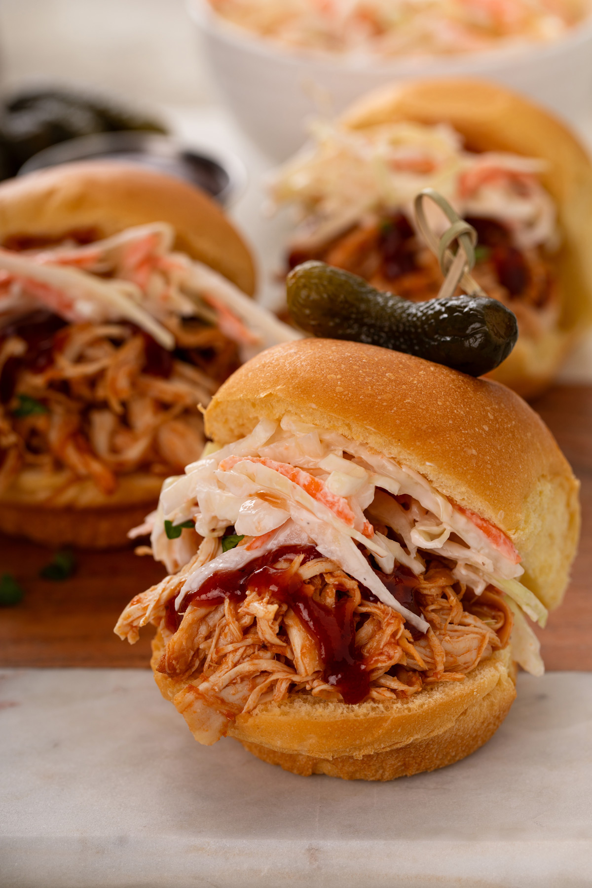 Close up of pulled chicken topped with slaw on a slider bun with a cornichon pickle on top.