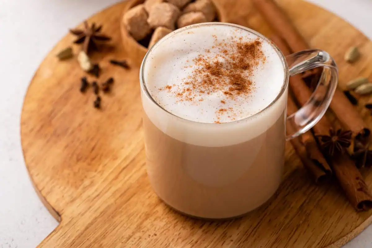 45-degree angle of a glass mug with a hot chai latte, sprinkled with cinnamon, set on a wooden board with whole spices.