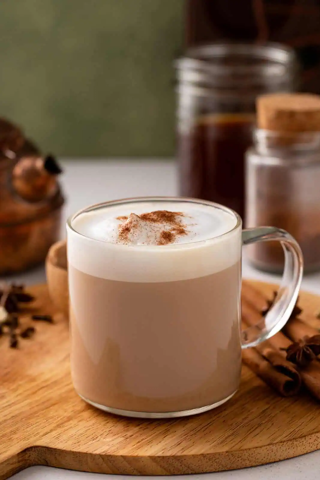 Glass mug filled with a hot chai latte and garnished with cinnamon set on a wooden board, with whole spices in the background.