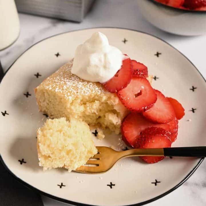 Plated slice of yellow cake topped with whipped cream and strawberries. A fork with a bite of the cake on it is on the plate.