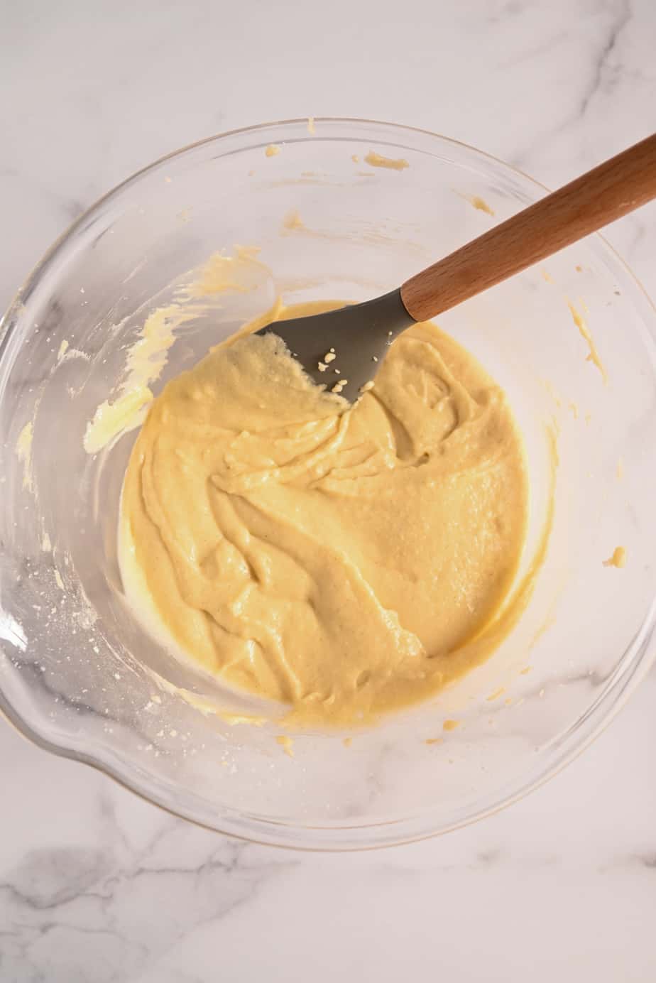 Yellow cake batter being stirred in a glass mixing bowl with a gray spatula.