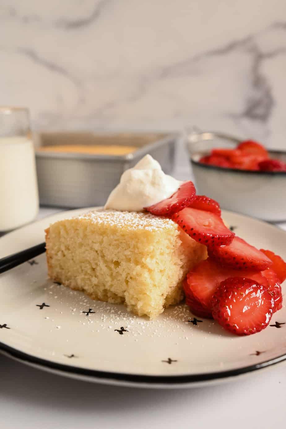 Slice of homemade yellow cake on a white and black plate, topped with strawberries and whipped cream.