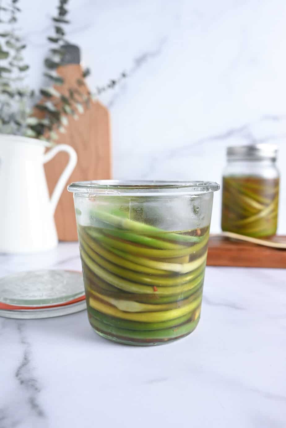 Opened glass jar of pickled garlic scapes set on a marble countertop.