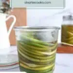 Opened glass jar of pickled garlic scapes set on a marble countertop. Text overlay includes recipe name.