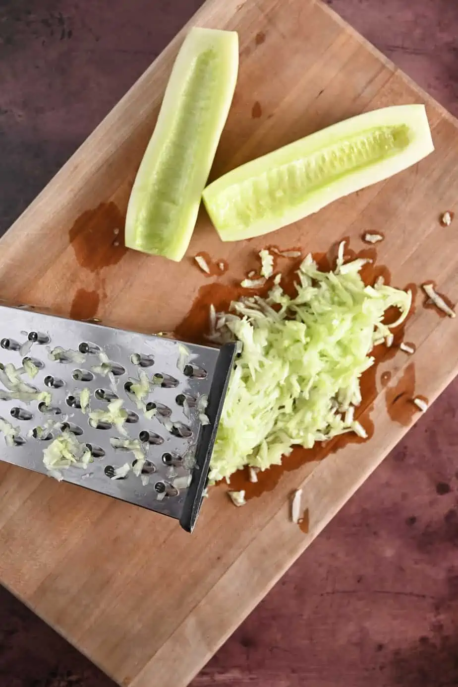 Box grater and grated cucumber on a wooden cutting board next to a halved and seeded cucumber.