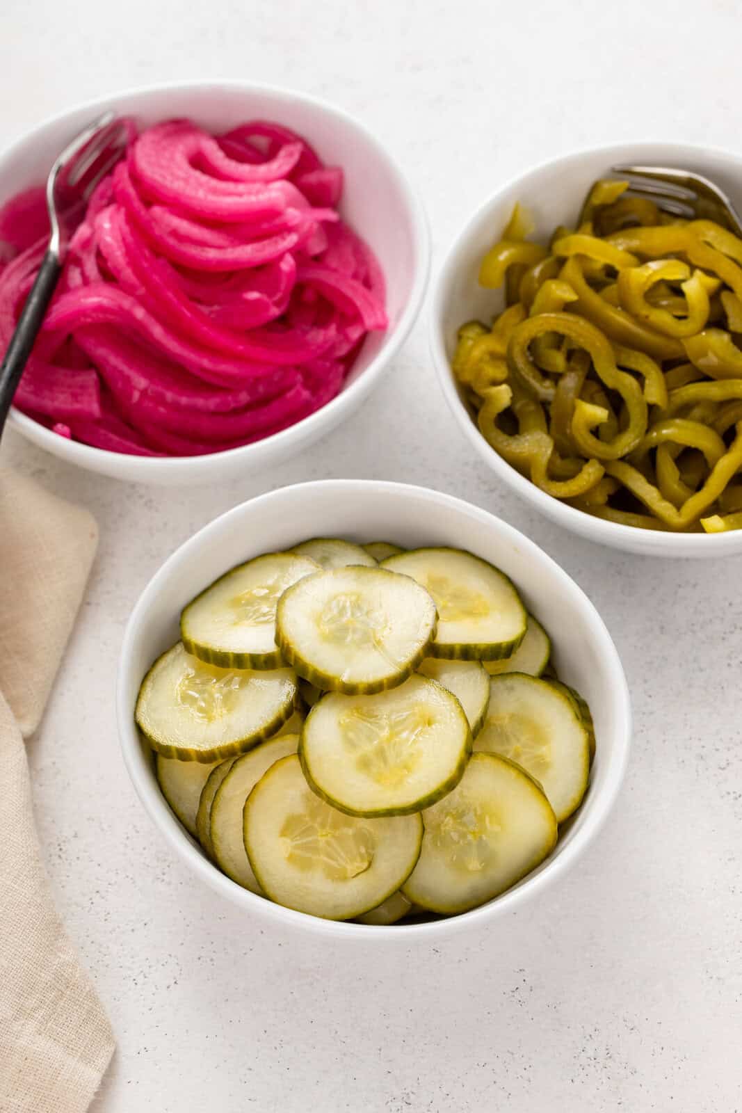 Three white bowls filled with pickled cucumbers, pickled red onion, and pickled green peppers.