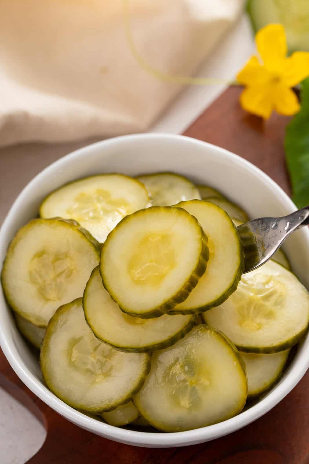 Fork lifting some slices of pickled cucumber out of a white bowl.