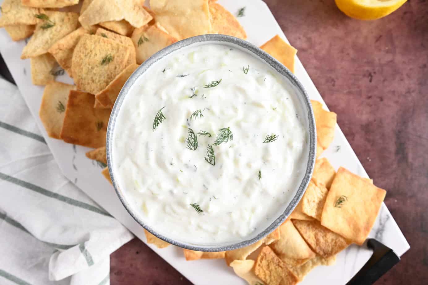 Bowl filled with homemade tzatziki surrounded by pita chips on a marble platter.