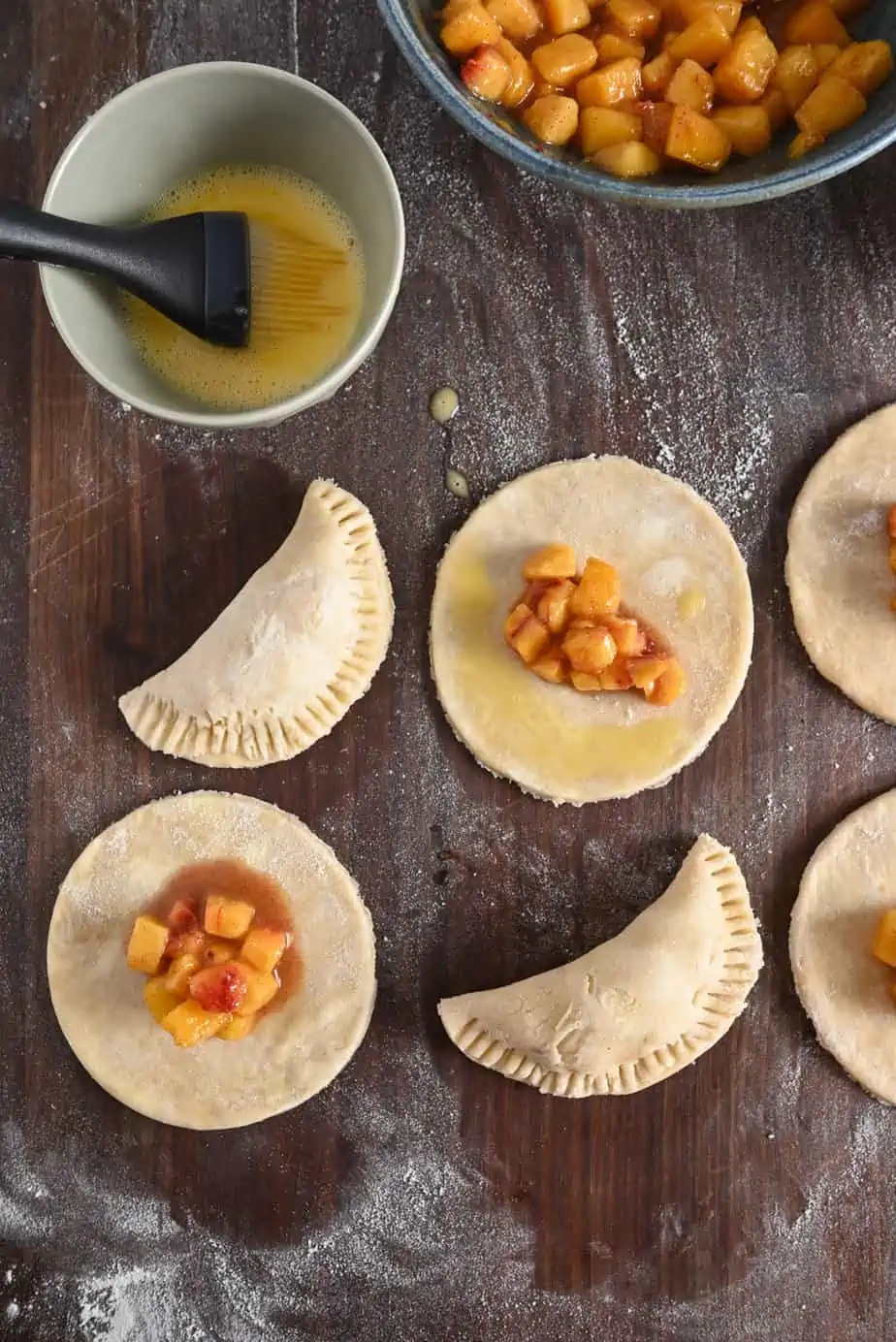 Egg wash being added to the edges of peach hand pies. Two of the hand pies are folded in half and crimped around the edges.