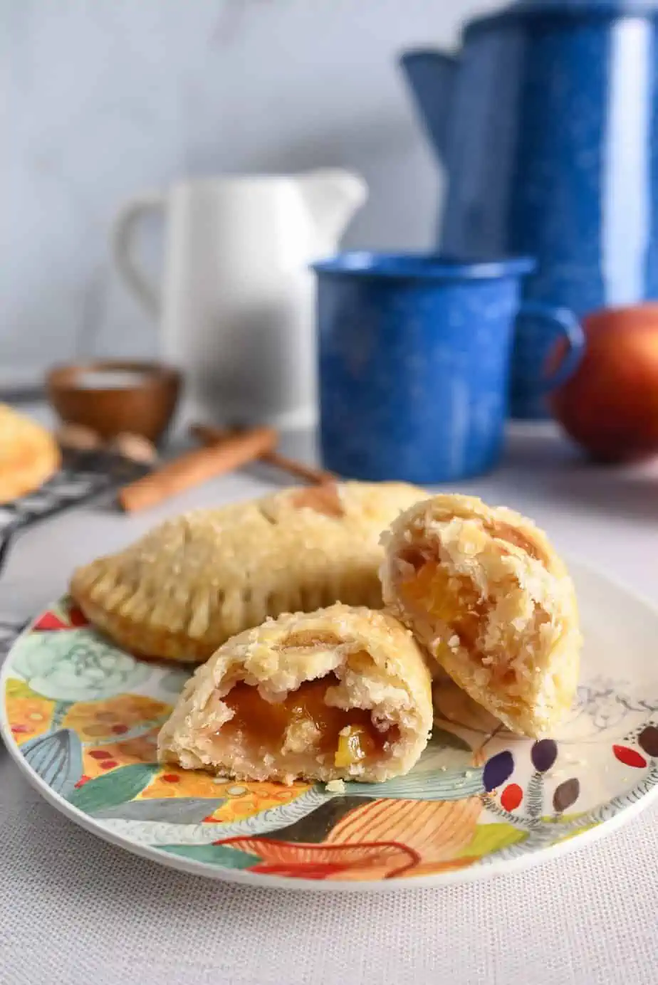 Two peach hand pies on a colorful plate. One of the pies is cut in half to show the peach filling. A coffee pot and coffee mug can be seen in the background.