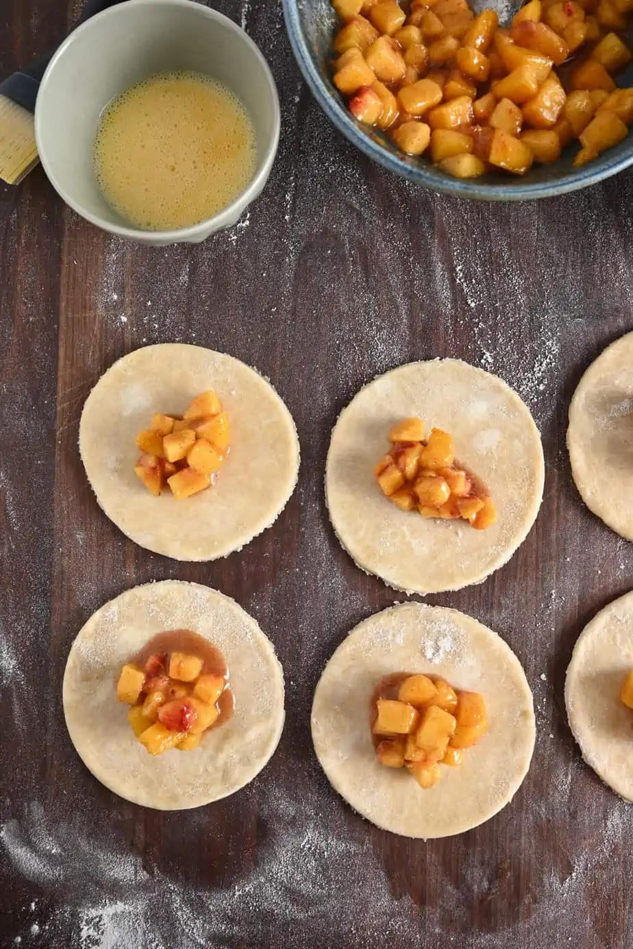 Pie crust circles, each with peach filling in the center, on a floured wooden board.