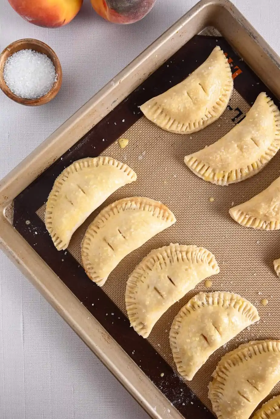 Shaped, crimped, and egg washed peach hand pies on a lined baking sheet, ready to go in the oven.