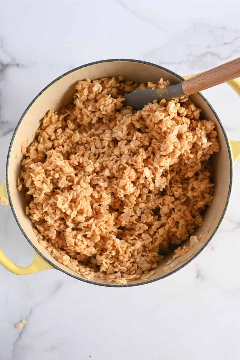 Mixture for peanut butter rice krispie bars being stirred with a spatula in a large yellow dutch oven.
