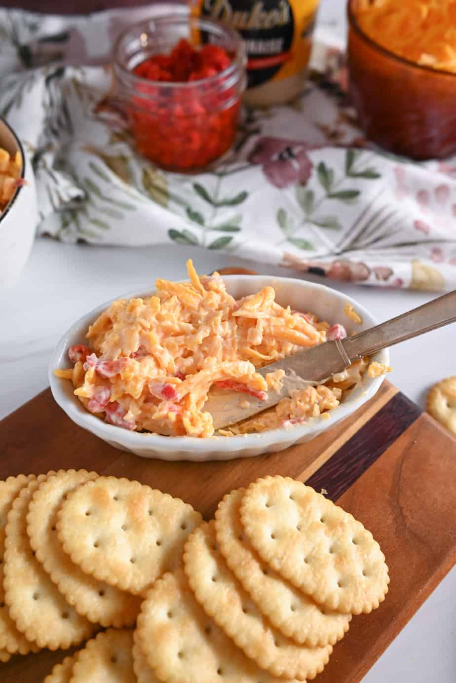 Butter knife in a small white dish filled with pimento cheese set on a wooden board with crackers.