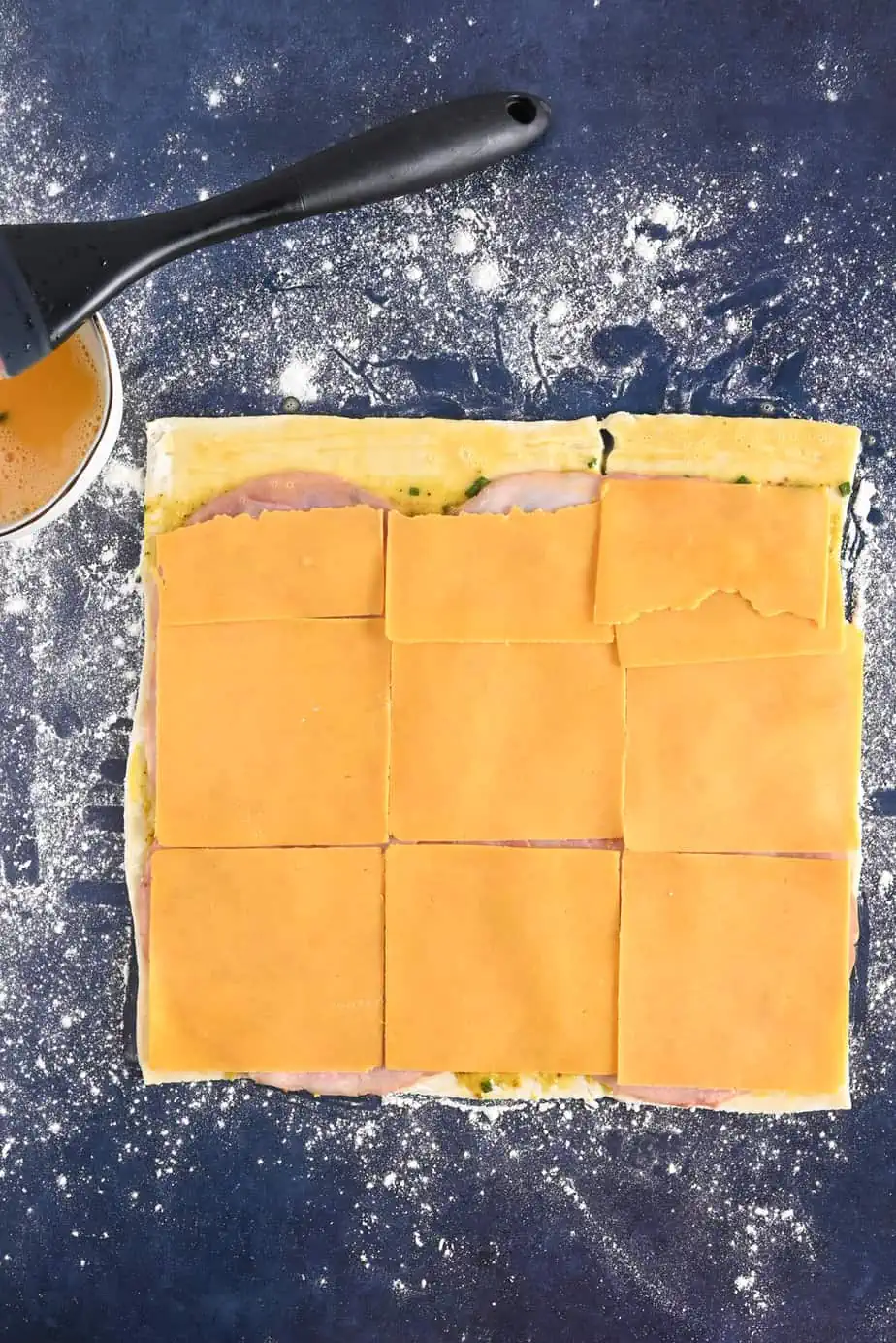 Ham and cheese layered on top of a sheet of puff pastry on a floured countertop.