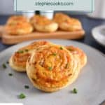 Three hot ham and cheese pinwheels on a gray plate with a platter of more pinwheels in the background. Text overlay includes recipe name.