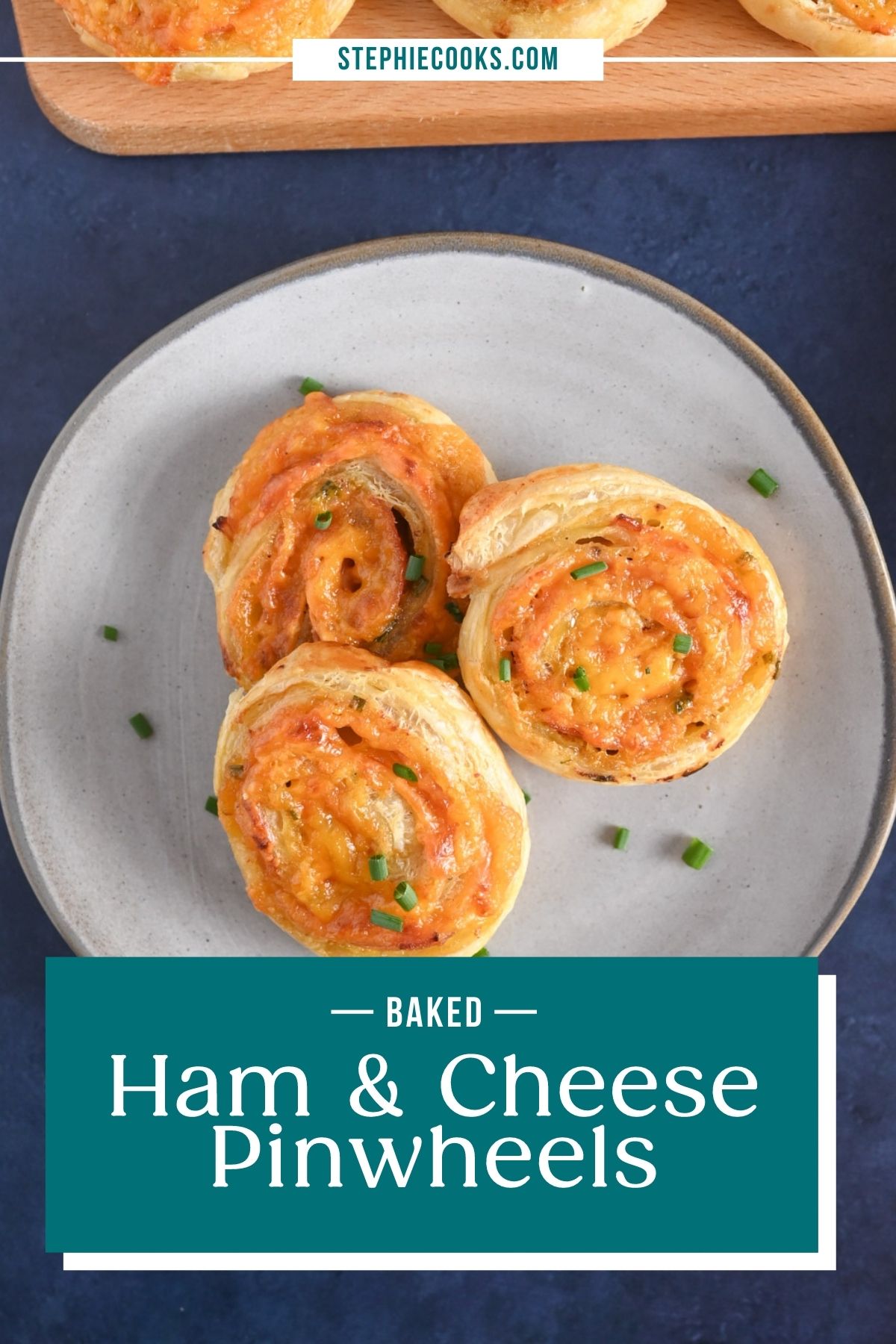 Overhead view of a gray plate holding three hot ham and cheese pinwheels set next to a wooden board holding more pinwheels. Text overlay includes recipe name.