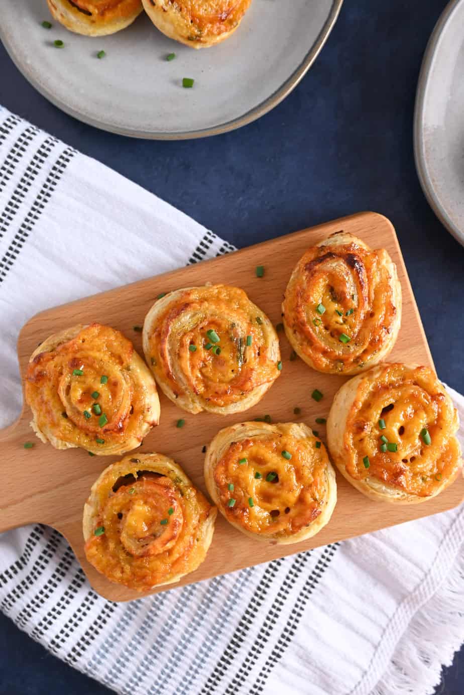 Wooden serving board holding hot ham and cheese pinwheels, surrounded by plates.