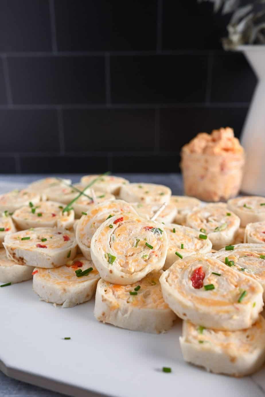 White tile platter piled with pimento cheese pinwheels. A bowl of pimento cheese is visible in the background.