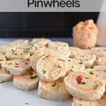 White tile platter piled with pimento cheese pinwheels. A bowl of pimento cheese is visible in the background. Text overlay includes recipe name.