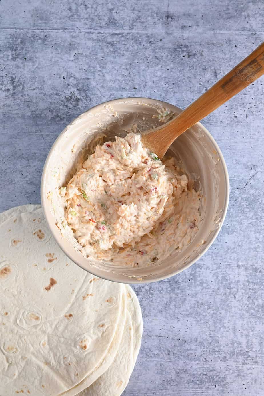 Pimento cheese pinwheel filling being stirred with a wooden spoon in a ceramic mixing bowl.