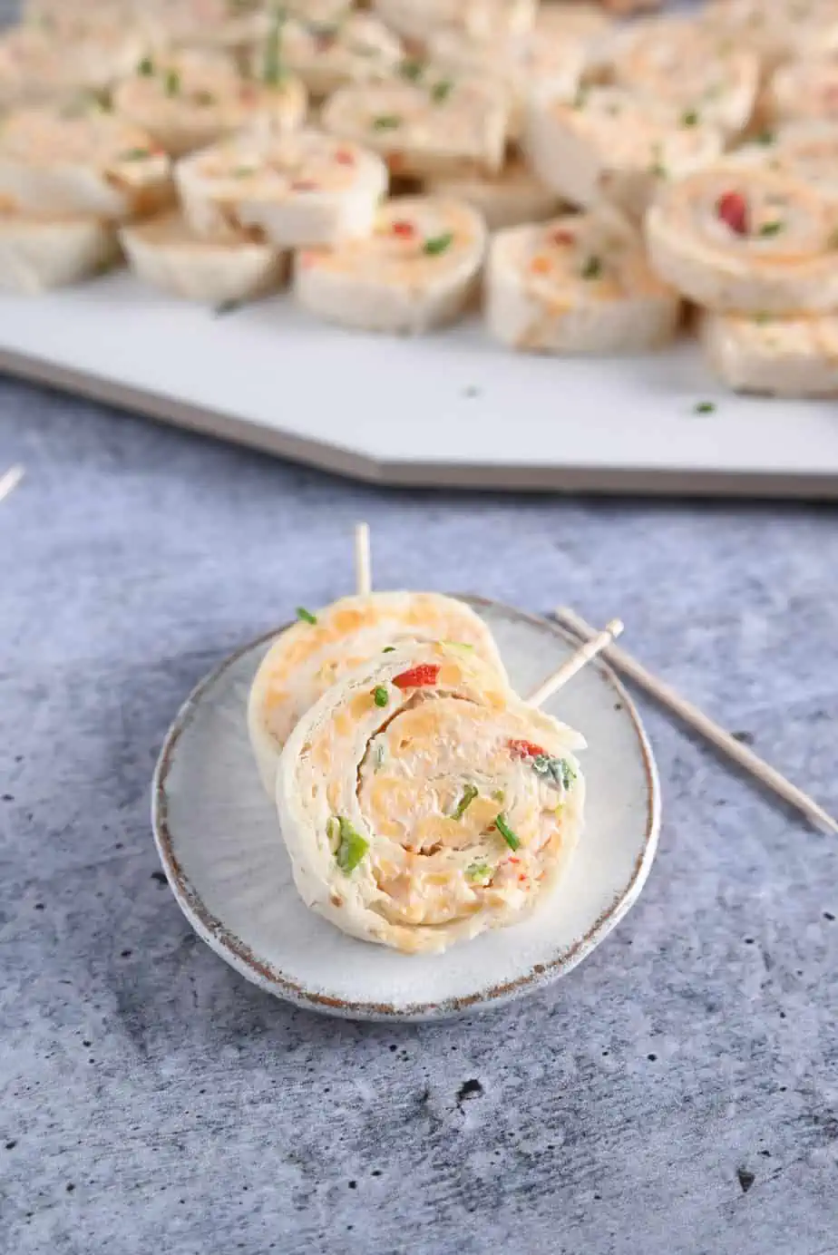 Two pimento cheese pinwheels on a small ceramic plate. A platter of the pinwheels is visible in the background.