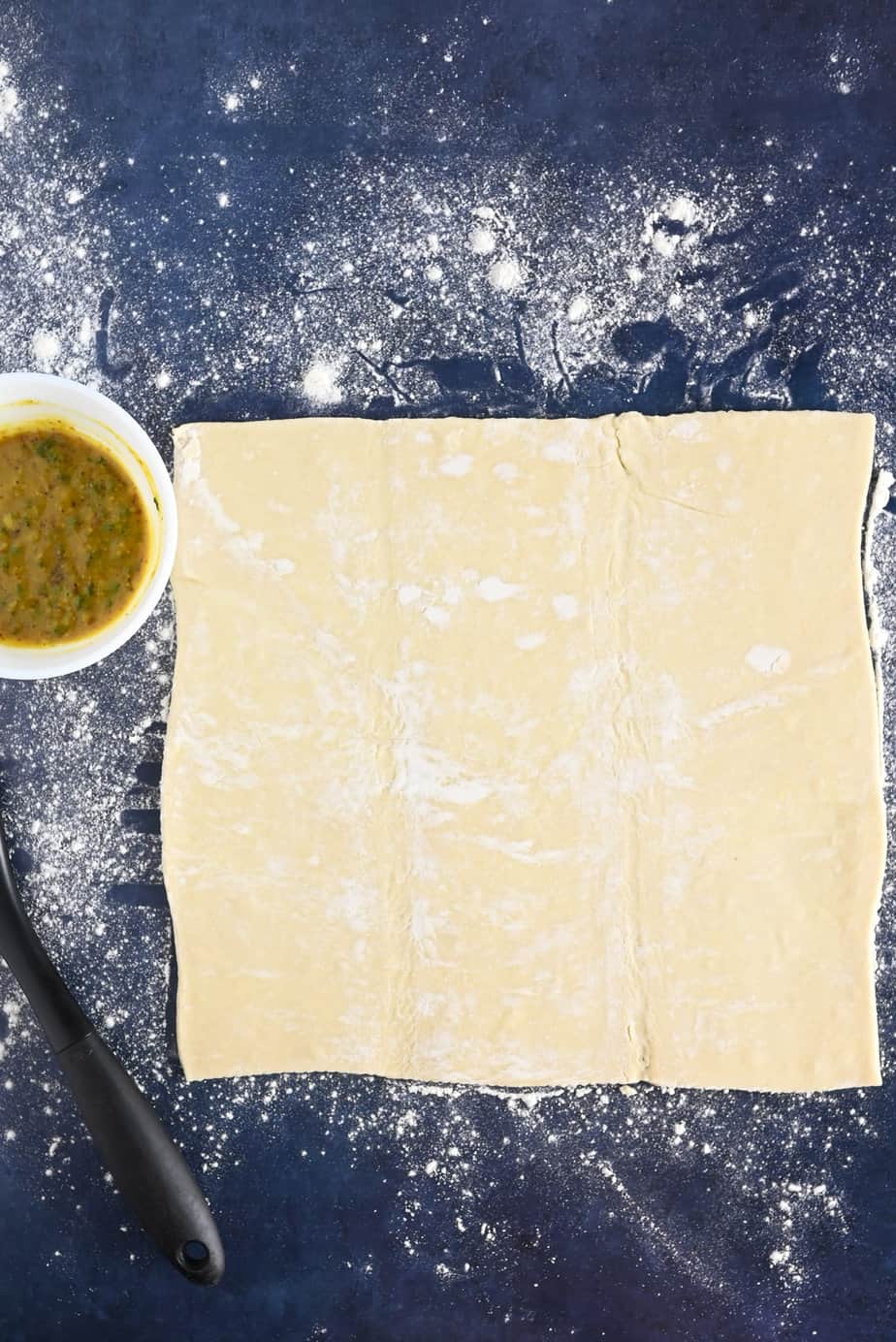 Sheet of puff pastry rolled out on a floured countertop next to a bowl of honey mustard spread.