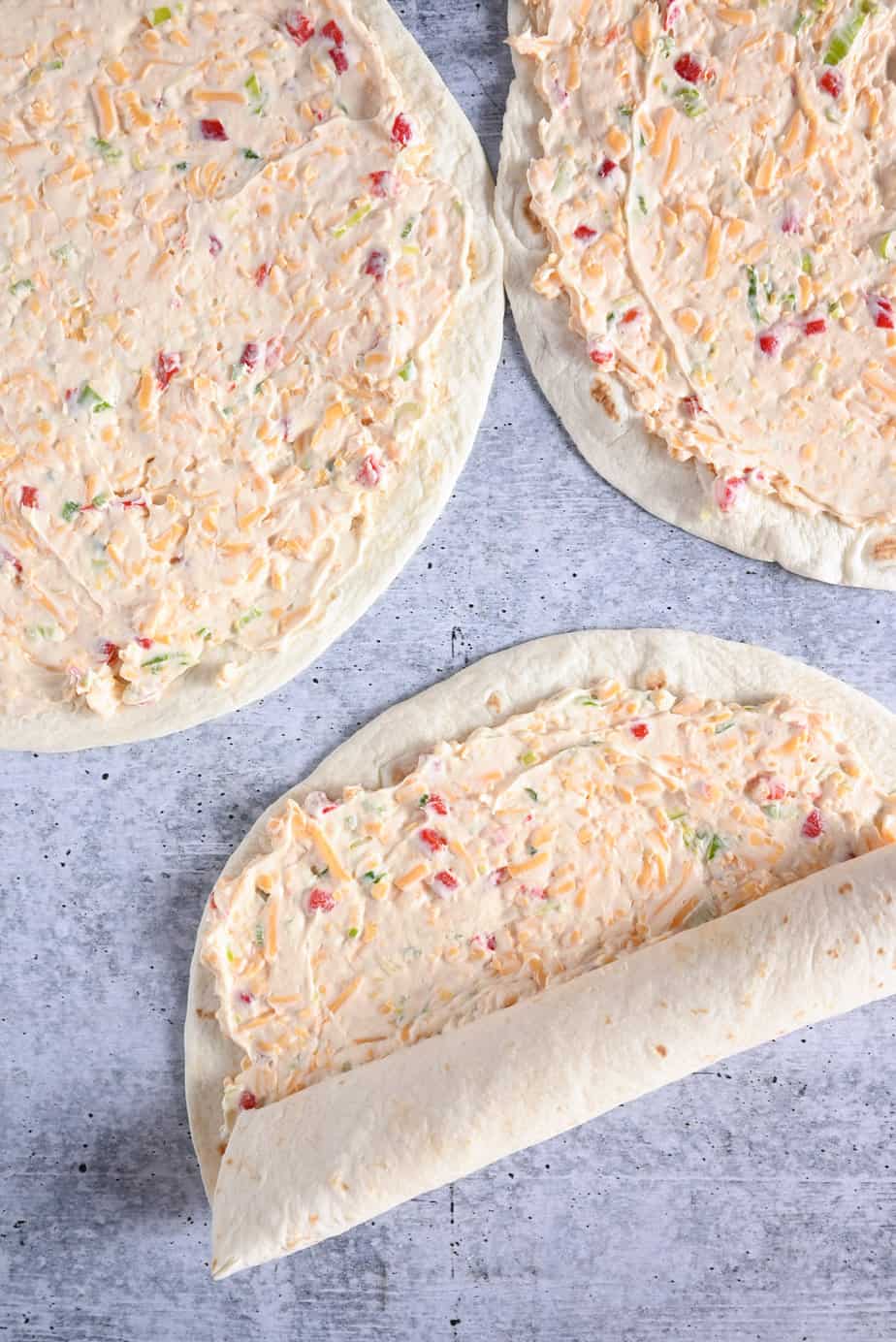 Flour tortillas spread with pimento cheese filling. One of the tortillas is halfway rolled up.