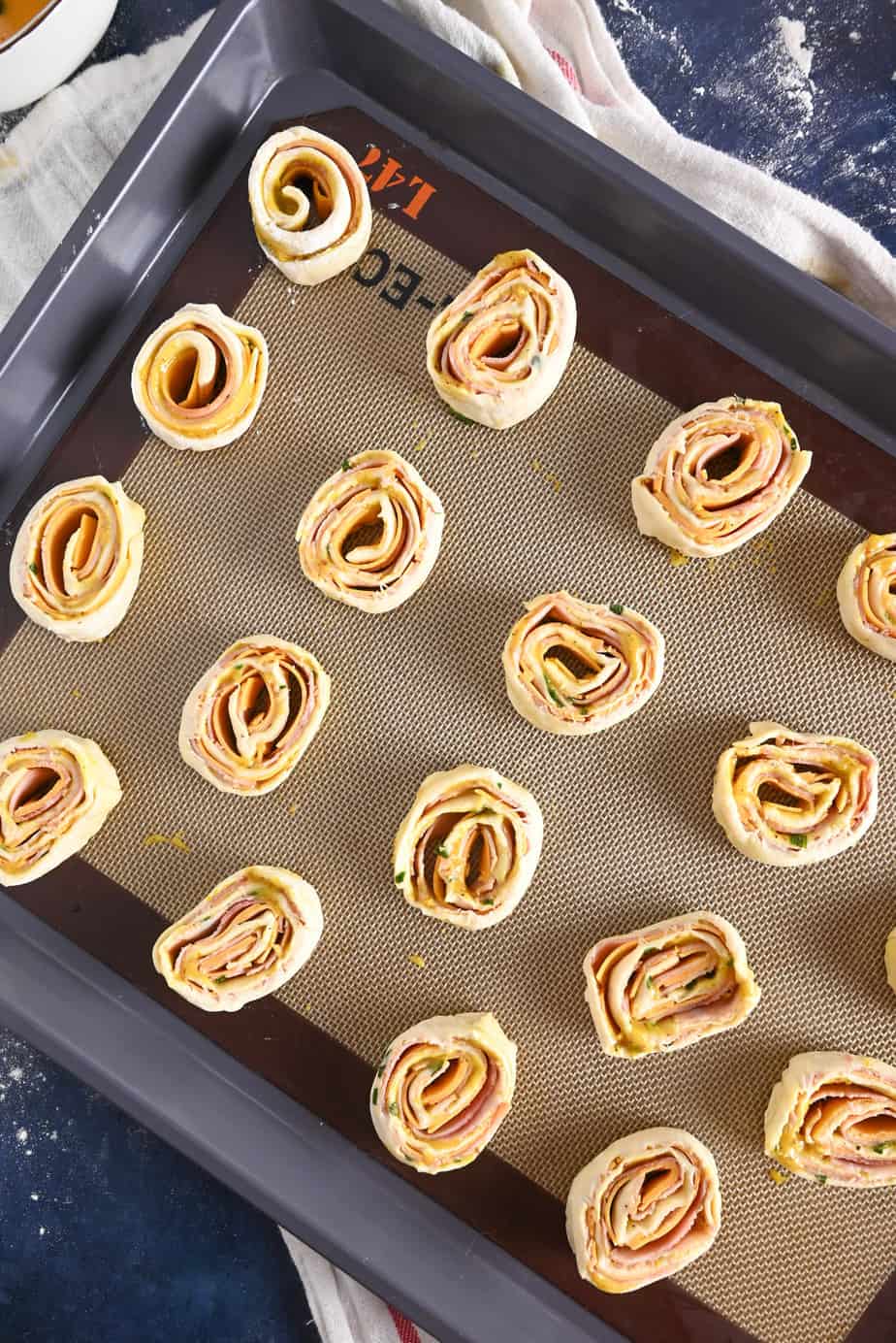 Unbaked hot ham and cheese pinwheels set on a baking sheet lined with a silicone baking mat, ready to go in the oven.