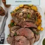 Overhead view of sliced roasted boneless leg of lamb on a platter, surrounded by roasted potatoes. Text overlay includes recipe name.