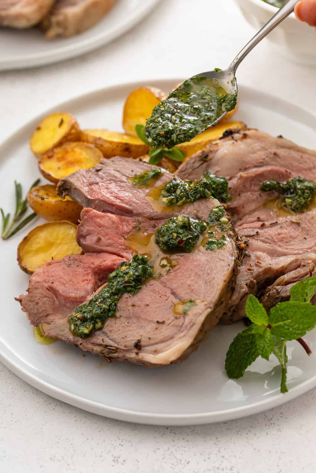 Mint chimichurri being spooned over slices of roasted boneless leg of lamb on a white plate.