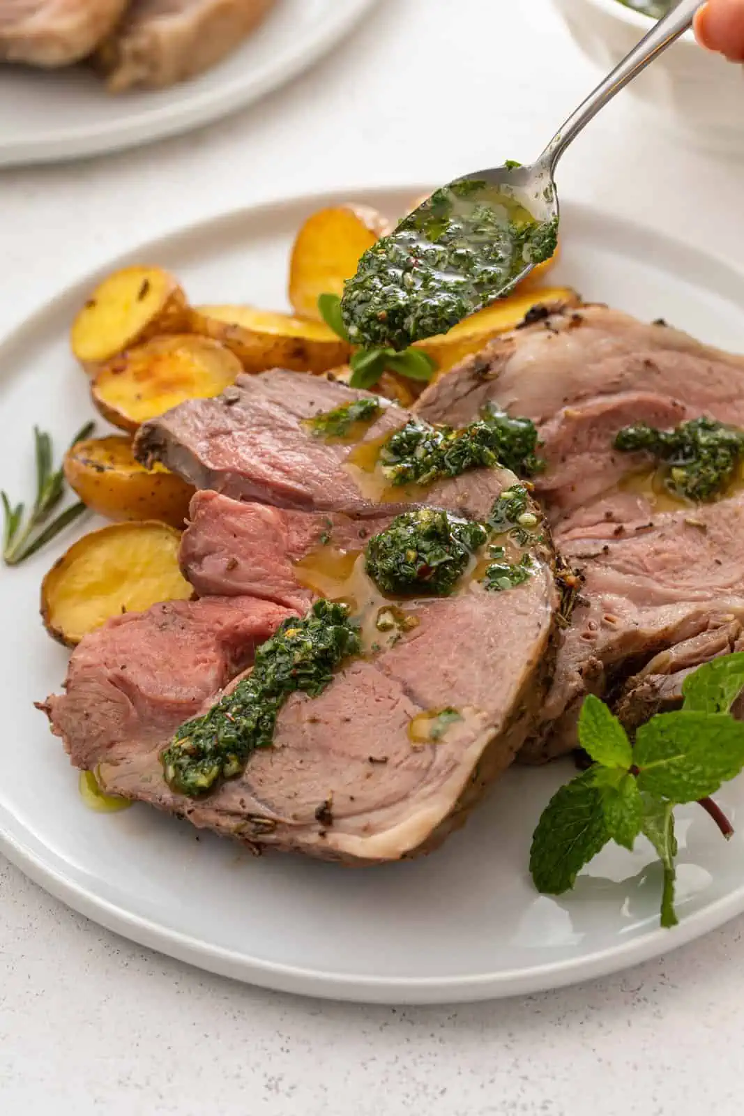 Mint chimichurri being spooned over slices of roasted boneless leg of lamb on a white plate.