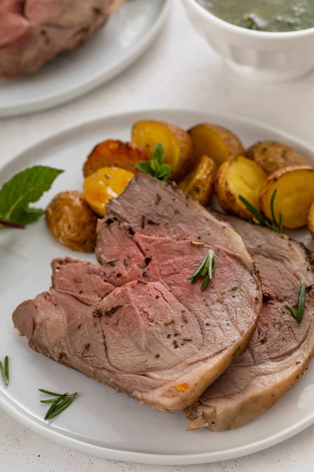 Two slices of boneless leg of lamb on a white plate next to roast potatoes.