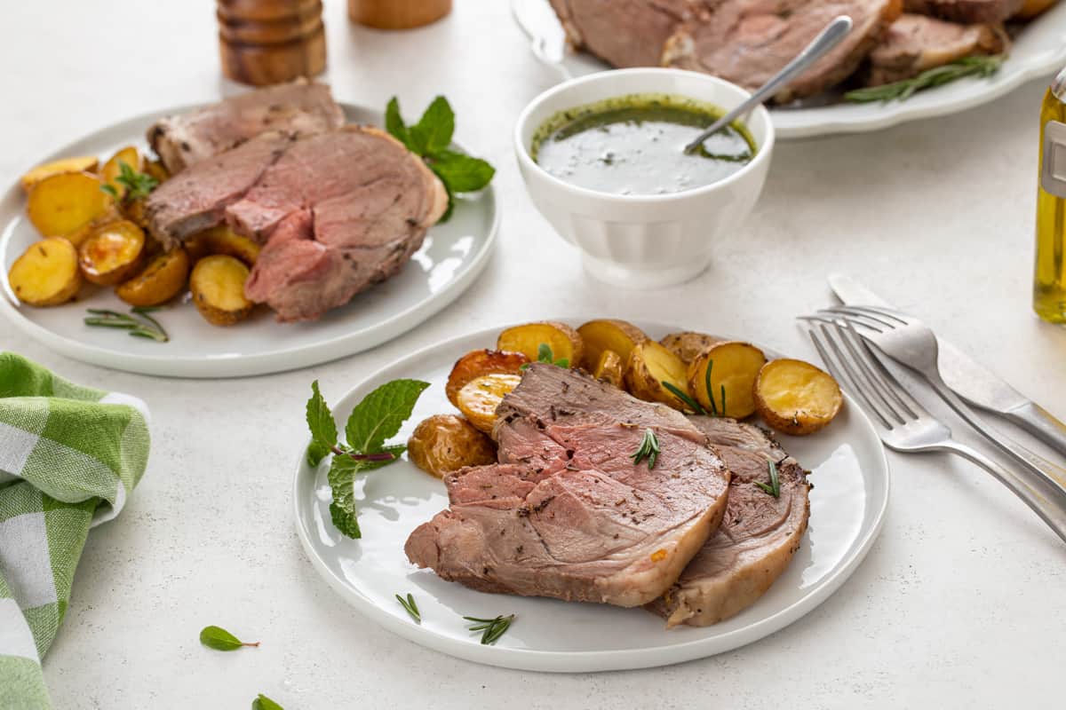 Two white plates, each holding slices of roasted boneless leg of lamb and potatoes with a bowl of mint chimichurri in the background.