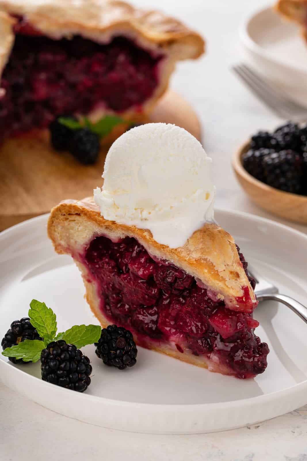 White plate holding a slice of blackberry pie topped with a scoop of vanilla ice cream.