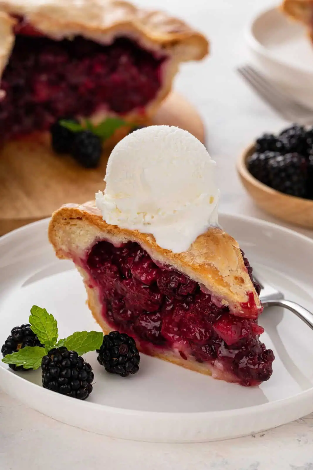 White plate holding a slice of blackberry pie topped with a scoop of vanilla ice cream.