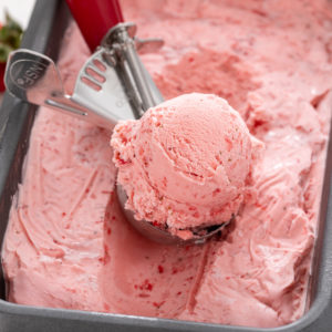 Ice cream scoop scooping up strawberry ice cream from a loaf pan container.