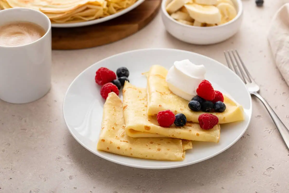Three folded crepes arranged on a white plate and topped with berries and whipped cream.