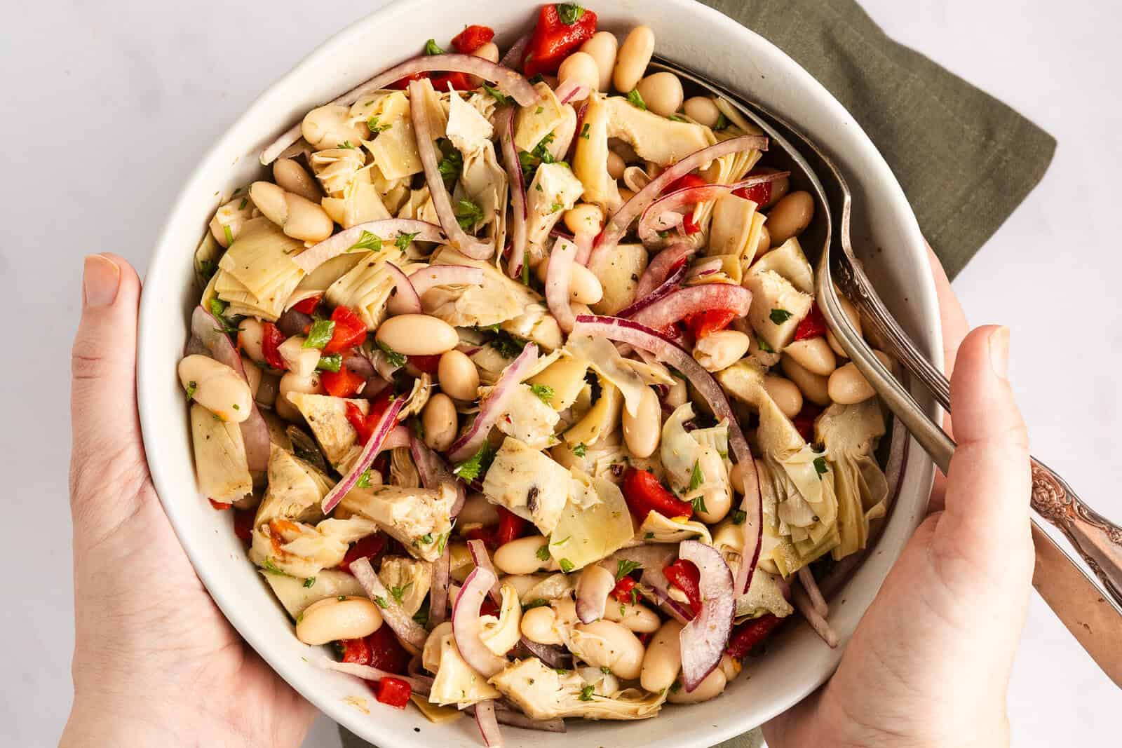 Overhead view of person holding a white serving bowl filled with white bean and artichoke salad.